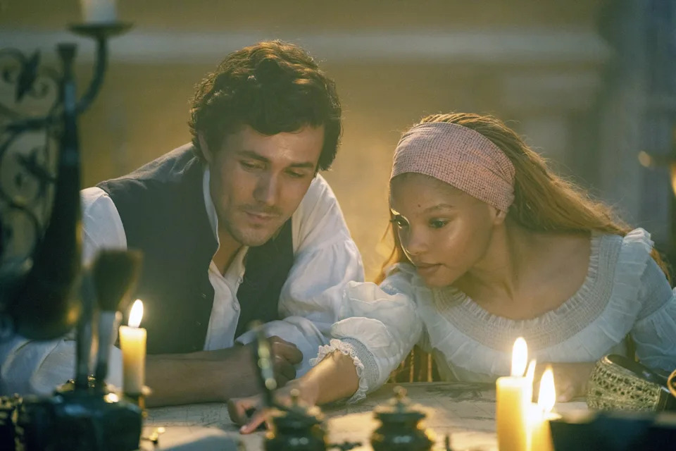 Two people sit closely at a candlelit table, engaging in quiet conversation. The woman wears a headband and ruffled dress. The scene is intimate