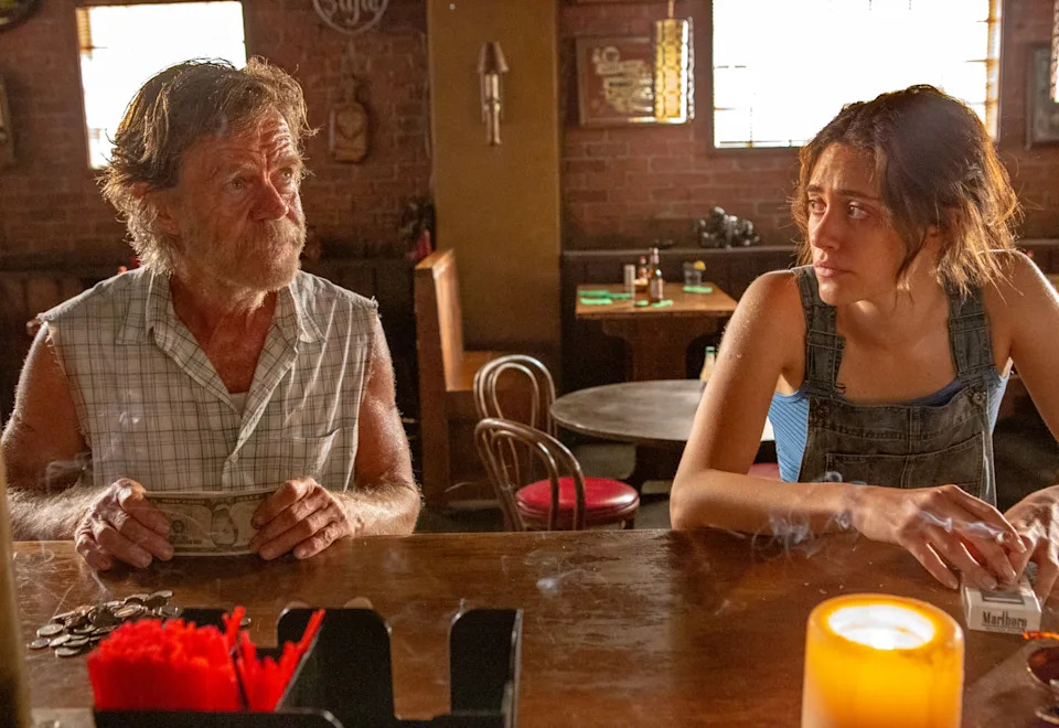 A man and a woman sit in a dimly lit bar. The man wears a sleeveless shirt; the woman has overalls on. Both have expressions of concern