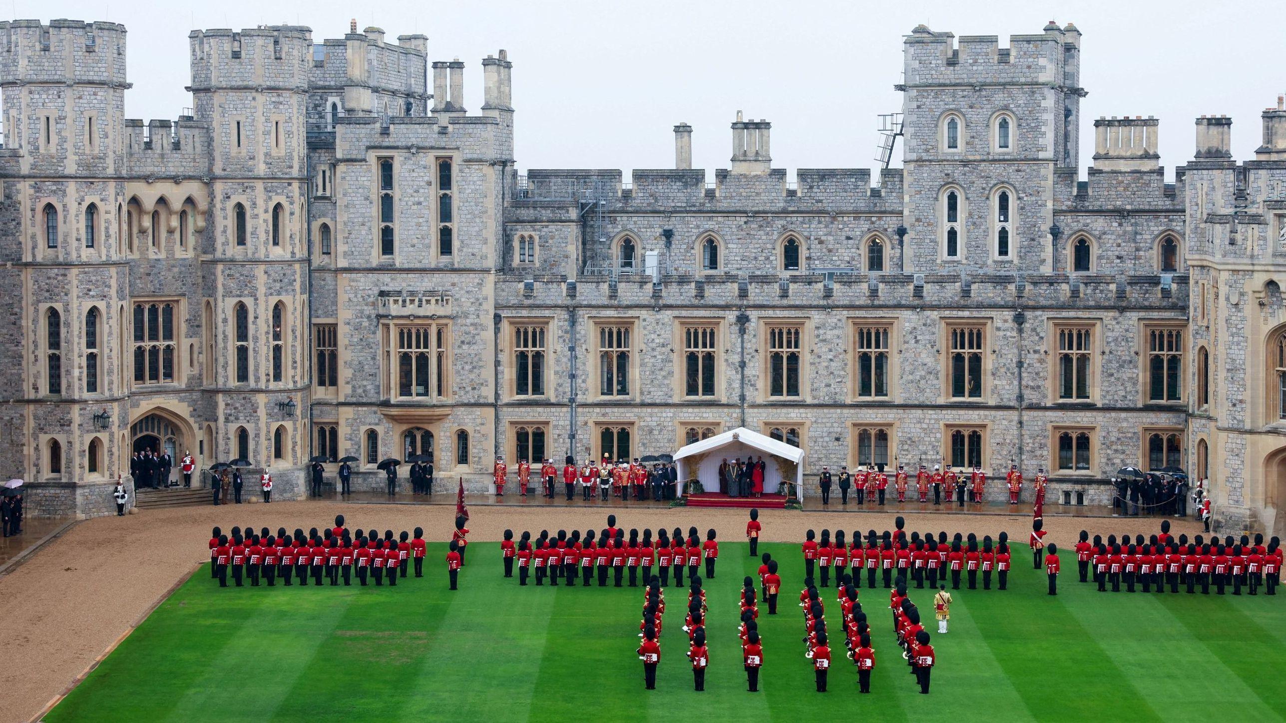 Windsor Castle with a large green lawn in the foreground, where troops wearing traditional red uniforms stand in formation