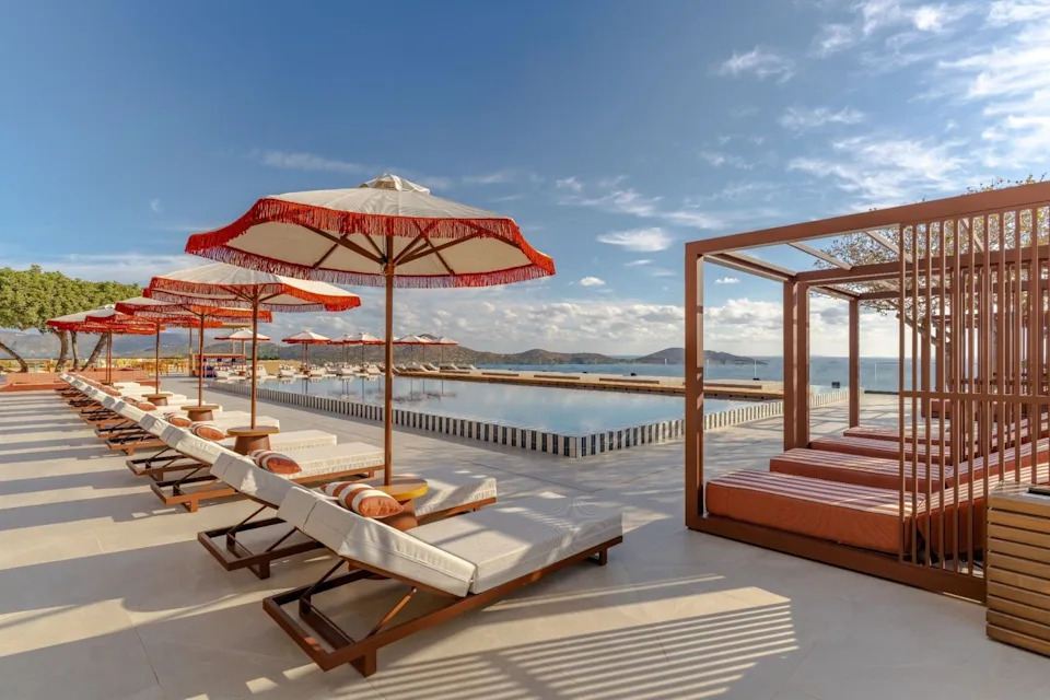 Poolside view with lounge chairs and umbrellas near a serene body of water. Clear sky and mountains in the background