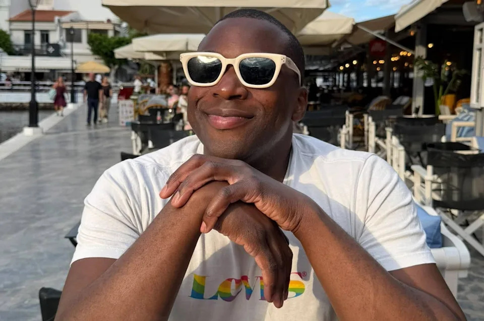 Person smiling at an outdoor café wearing sunglasses and a T-shirt with "Love" in rainbow print