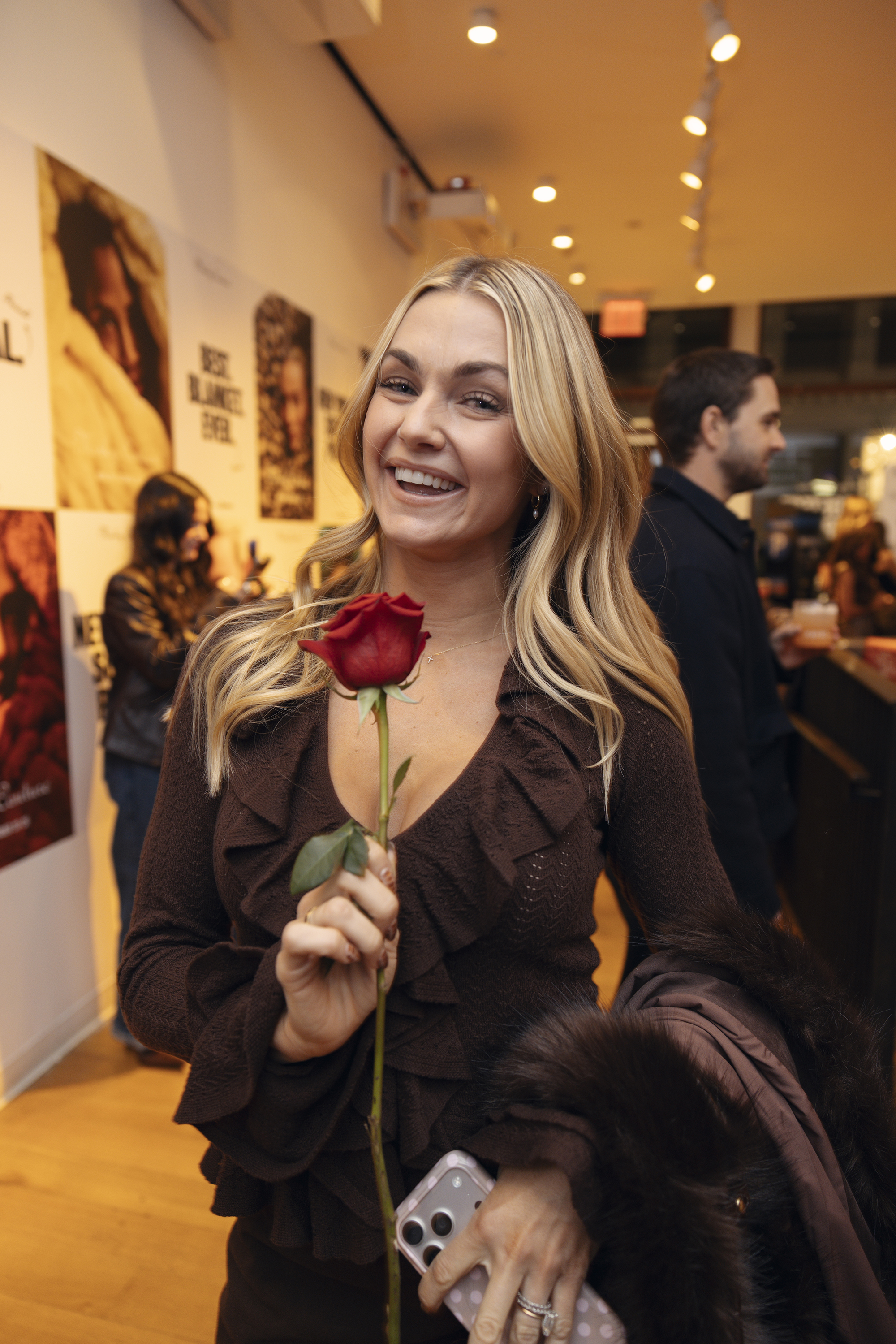 A smiling blond woman holds a red rose in an indoor setting.