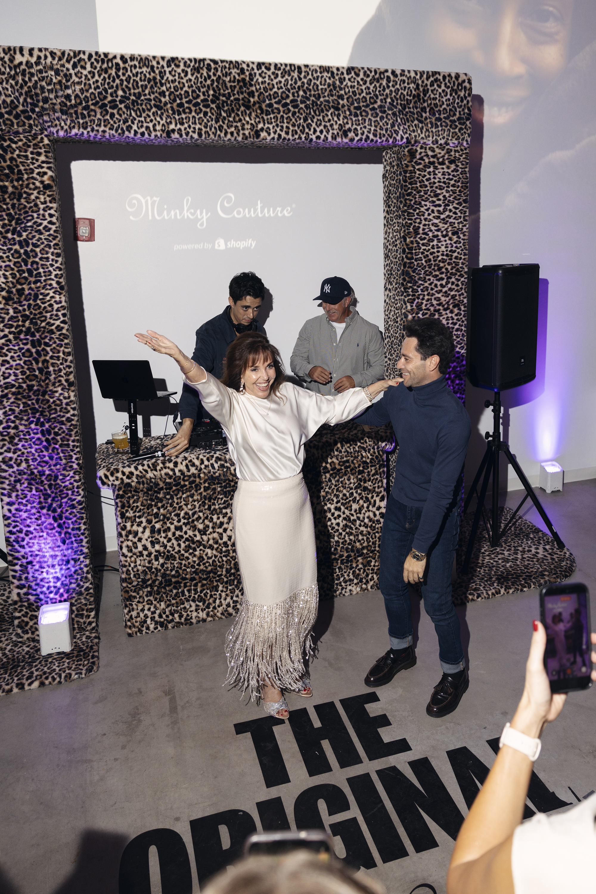 A woman in a white top and fringed skirt dances with a man in a blue turtleneck in front of a leopard-print DJ booth.