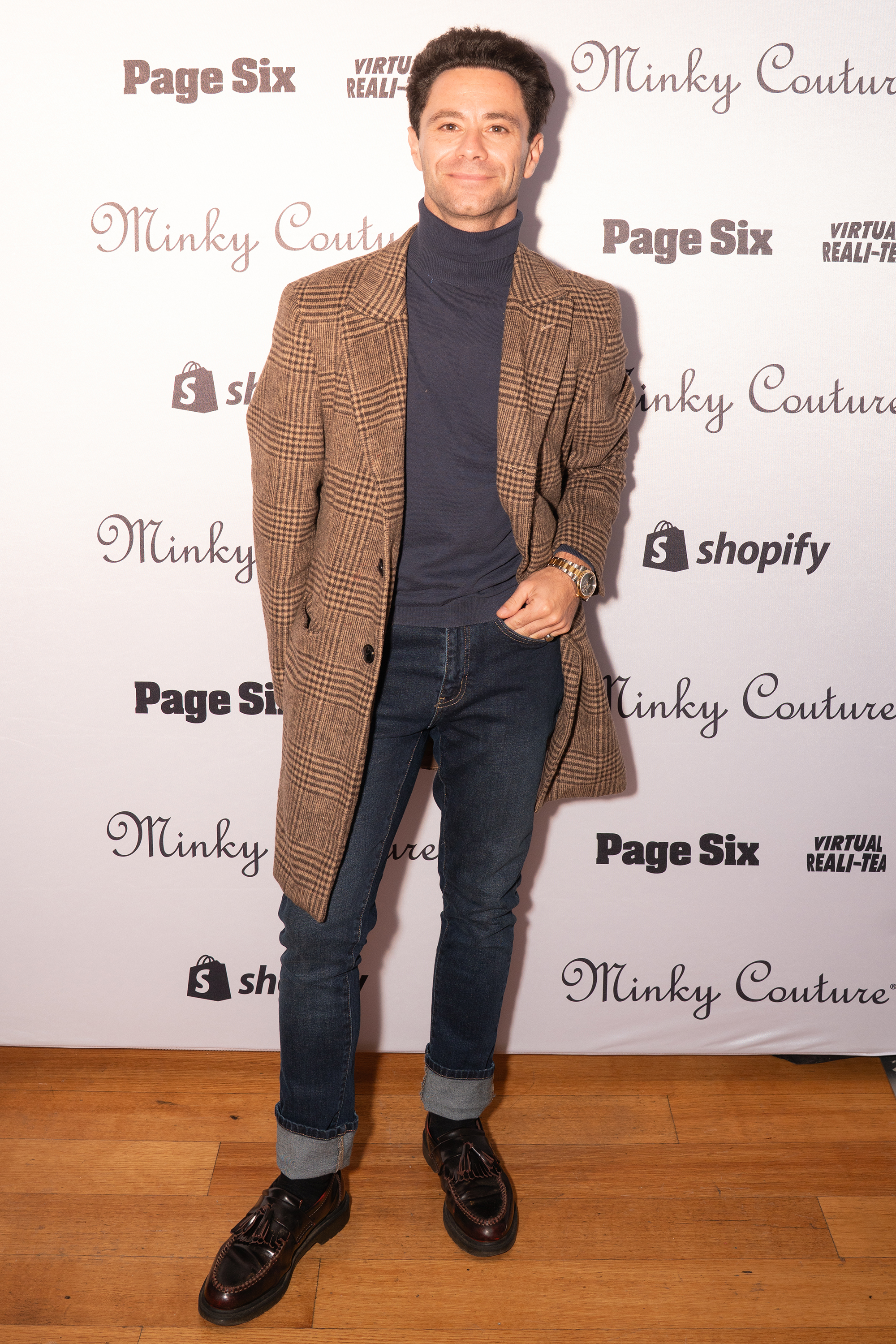 A man wearing a brown plaid coat, blue turtleneck, and dark jeans stands in front of a branded step-and-repeat backdrop.