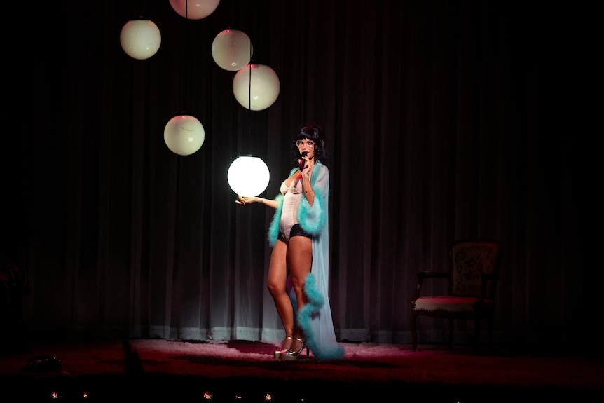 Lily Allen, wearing a blue dressing gown, stands beside several round light sources on stage.