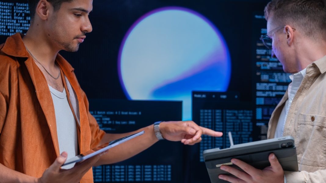 AI is killing off entry-level cybersecurity jobs | Information Age Two young male cybersecurity workers holding tablets and working in front of a screen displaying computer code.
