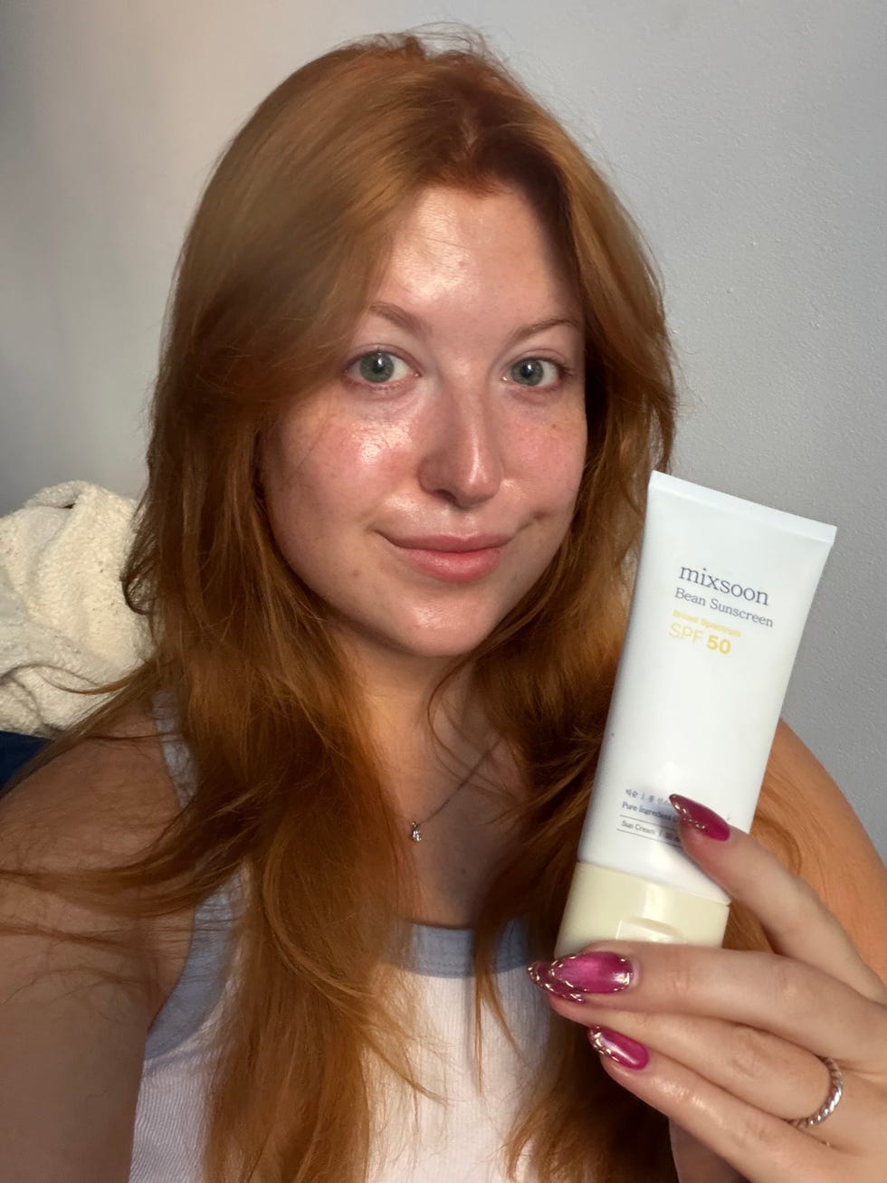 Person with long red hair holding a sunscreen tube, smiling at the camera.